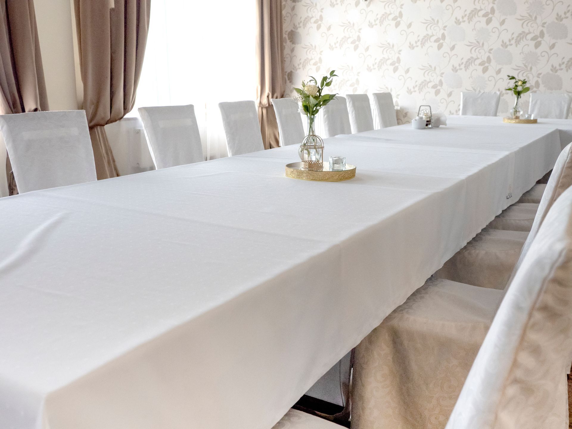 A long table covered with a white tablecloth, set with chairs and floral centerpieces in a bright, formal event room.