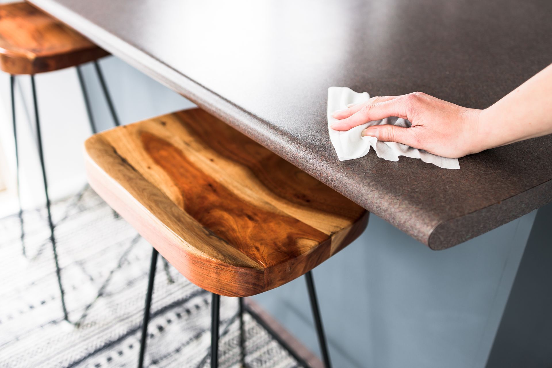 A hand wipes a dark, textured kitchen countertop with a white cloth above a wooden stool with black metal legs.